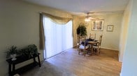 This single-floor residence offers a comfortable and well-lit living environment with a thoughtful layout. The dining area is a standout feature, showcasing light hardwood flooring that adds warmth and a clean aesthetic to the space. A large sliding glass door, dressed with elegant vertical blinds and a decorative gold valance, allows an abundance of natural light to fill the room while providing direct access to the outdoors. A ceiling fan with an integrated light fixture is centrally positioned overhead, contributing to both comfort and ambiance. The dining area accommodates a wooden table and chair set, complemented by decorative greenery and framed wall art that enhance the inviting atmosphere. Adjacent carpeted areas suggest a seamless transition into additional living spaces, adding variety in flooring and texture throughout the home. The warm, neutral wall tones create a cohesive backdrop that ties the interior together. Overall, this residence combines functional design with comfortable, everyday livability, making it a practical and pleasant space suitable for a variety of lifestyles.