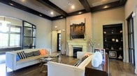 This thoughtfully designed single-floor space makes a strong first impression with its refined architectural details and contemporary aesthetic. The main living area is defined by dramatic dark wood coffered beams that contrast beautifully against the light, neutral ceiling, adding both structure and visual depth to the room. Elevated ceilings amplify the sense of openness and grandeur throughout.

A standout feature of the space is the elegant fireplace, framed by a classic white stone mantel and flanked by decorative accents, anchoring the room as its natural focal point. Above it, a large-scale abstract artwork adds a bold artistic statement that complements the neutral color palette of the walls.

The flooring features rich dark hardwood, grounding the space with warmth and sophistication. Comfortable cream-toned sofas accented with black-and-white patterned and coral throw pillows contribute to a polished yet inviting atmosphere. A sleek glass-and-gold coffee table adds a modern touch to the seating arrangement.

Recessed lighting throughout ensures the space is well-illuminated, while large dark-framed windows invite natural light and hint at a strong indoor-outdoor connection. Overall, this space balances contemporary design with classic architectural elements, creating an environment that is both functional and visually impressive.