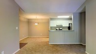 This single-floor residence offers an open-concept layout that seamlessly connects the kitchen and living areas, creating a spacious and functional environment. The kitchen is designed with clean white cabinetry, a tiled backsplash, and stainless steel appliances including a refrigerator and electric range, providing both style and practicality. A raised breakfast bar countertop serves as a natural divider between the kitchen and the adjoining living space, ideal for casual dining or entertaining. The living area is finished with wall-to-wall neutral carpeting that adds warmth and comfort underfoot. Soft, neutral gray walls throughout create a calm and cohesive atmosphere, offering a versatile backdrop for a variety of interior styles. A pendant light fixture with a classic bowl design hangs in the dining area, providing focused ambient lighting. Recessed and overhead fluorescent lighting in the kitchen ensures a well-lit workspace. The overall design is practical and understated, with clean lines and a muted color palette that gives the space a bright, airy feel. The layout maximizes the sense of openness across the single floor, making it both easy to navigate and comfortable for everyday living.