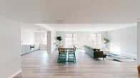 Located at 1435 51st St in North Bergen, NJ, this single-floor space offers a thoughtfully designed open-plan layout that seamlessly integrates a spacious living room and a contemporary kitchen. Light-toned hardwood flooring runs throughout, lending a clean, cohesive aesthetic to the entire floor.

The living area is generously proportioned and bathed in natural light from multiple large windows fitted with neutral roller shades, offering views of the surrounding greenery. Recessed lighting complements the natural light, while soft gray walls and a textured accent wall add visual depth. The space comfortably accommodates a large sectional sofa, a sizable dining table with seating for many, and a dedicated lounge corner, making it well-suited for both casual gatherings and collaborative use. Glass double doors at the rear provide a transparent connection to adjacent areas.

The kitchen is finished to a high standard, featuring shaker-style cabinetry in soft gray, elegant marble-look quartz countertops, and a full suite of stainless steel appliances including a side-by-side refrigerator with water dispenser, a dishwasher, and a built-in microwave. The overall ambiance of the space is modern, airy, and welcoming, ideal for community or professional use.