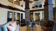 This impressive single-floor space makes an immediate impact with its dramatic double-height ceilings and open, airy layout. A commanding stacked stone column serves as the architectural centerpiece of the main lounge area, lending a rustic yet refined character to the space. Dark wood beam accents contrast beautifully against warm, neutral-toned walls, creating a rich and inviting atmosphere throughout.

The lounge area is thoughtfully furnished with sleek black leather chairs and a plush sofa accented with decorative pillows, all arranged around a metallic oval coffee table atop a woven area rug. Large equestrian-themed artwork displayed high on the upper walls adds a distinctive, curated touch to the interior design.

Recessed lighting and pendant fixtures illuminate the space, while the elevated ceiling cutouts add architectural depth and visual interest. Multiple corridors branch off from the main area, suggesting functional spaces beyond, including what appears to be a reception or service counter visible to one side. The polished concrete flooring ties the design elements together, completing a space that balances warmth and sophistication with an upscale, welcoming ambiance.