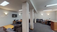 This single-floor space is thoughtfully designed to accommodate a variety of group activities and gatherings. The open layout is anchored by structural white columns that naturally define distinct functional zones without the need for full walls, creating a sense of flow throughout the interior.

The seating area features multiple sofas and upholstered chairs arranged around a central coffee table, providing a comfortable lounge setting. Adjacent to this is a dedicated meeting or dining area with several round wooden tables surrounded by rolling leather chairs, suitable for small group discussions or meals.

The fully equipped kitchen includes warm wood cabinetry, granite countertops, stainless steel appliances—including a range, over-the-range microwave, and refrigerator—and a tile floor. A peninsula counter extends into the space, offering additional workspace and a natural transition between the kitchen and the main gathering area.

Throughout, wall-mounted sconce lighting provides a warm, even ambiance complemented by recessed ceiling lights and flush fluorescent fixtures. The carpeted floors in the main area add comfort underfoot. A wall-mounted flat-screen television and multiple doorways leading to additional rooms complete this functional, community-oriented space.