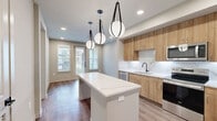 Located at 2080 FM 1102 in New Braunfels, TX, this thoughtfully designed one-bedroom, one-bathroom unit offers a modern aesthetic with high-quality finishes throughout. The open-concept kitchen serves as the heart of the space, featuring warm wood-toned cabinetry, a striking white quartz island with subtle veining, a marble-look tile backsplash, and stainless steel appliances including a French door refrigerator. Dramatic globe pendant lights hang above the island, adding a contemporary focal point, while recessed lighting and wood-look flooring carry throughout the main living areas. The bedroom is a comfortable, well-lit retreat with two windows that invite natural light and offer views of the surrounding community. A ceiling fan adds both function and style to the room. The bathroom is elegantly appointed with a wood-tone vanity, white quartz countertop, matte black hardware, large-format tile flooring, a backlit mirror, and a glass-enclosed shower with marble-style tile surround. Directly accessible from the bathroom, the walk-in closet features built-in shelving for organized storage. A hallway entry area includes additional built-in shelving, rounding out this well-planned and modern living space.