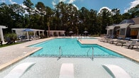 Located at 1630 Lewis Creek Dr in Wilmington, North Carolina, this outdoor community amenity space is designed to deliver a resort-style experience for its residents. At its centerpiece is a generously sized, multi-section swimming pool with crystal-clear water, accessible via stainless steel handrails and steps. The surrounding pool deck is finished in textured concrete and furnished with chaise lounges and shaded umbrella seating, all set against a lush backdrop of tall Carolina pines and palm trees that lend a coastal Southern character to the environment.

Adjacent to the pool is a covered pavilion that serves as the social hub of the outdoor space. The pavilion features a vaulted wood-paneled ceiling, brick-accented columns, and an open-air design that invites natural breezes and garden views. It is equipped with an outdoor dining table and chairs, comfortable lounge seating with accent pillows, a wall-mounted television, and a nearby grilling station — making it well-suited for both casual gatherings and larger community events. The polished aggregate concrete flooring ties the indoor and outdoor areas together seamlessly. A white pergola structure visible in the background adds additional shaded outdoor seating, completing this well-appointed community retreat.