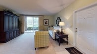 This single-floor residence offers a thoughtfully designed layout with a cohesive neutral palette throughout. The kitchen is configured as a galley-style space, featuring white cabinetry, white appliances including a full-size refrigerator, a built-in microwave, a double sink, and a cooktop. Light wood-toned flooring runs through the kitchen and connects to an adjacent hallway, enhancing the sense of flow.

The living room is a welcoming, carpeted space bathed in natural light from a large sliding glass door that opens to an outdoor area with lush greenery and palm tree views. Soft sage-green walls and a neutral ceiling complement the comfortable furnishings, creating a relaxed yet polished atmosphere.

The bedrooms are carpeted and feature textured ceilings, neutral walls, and elegant dark wood four-poster bed frames. One bedroom includes a generously sized window with outdoor views of mature trees, adding a serene, natural quality to the space.

The bathroom is clean and practical, offering a single vanity with a large mirror, a toilet, and a shower-tub combination enclosed by a curtain. Tile flooring and light-toned walls maintain a bright, functional character consistent with the rest of the residence.