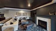 This single-floor space is thoughtfully designed as a premium entertainment and social hub, combining refined aesthetics with practical amenities across several distinct zones. The kitchenette is a standout feature, showcasing rich dark cabinetry with gold hardware, sweeping granite countertops with striking veining, and a bold deep-blue subway tile backsplash. A stainless steel sink, built-in microwave, wall oven, and refrigerator complete the setup, while industrial-style pendant lights add character overhead. Wood-look plank flooring runs throughout, adding warmth and visual continuity. The lounge area exudes an intimate, upscale ambiance with a dramatic dark ceiling framed by rich crown molding, a herringbone tile fireplace flanked by custom dark cabinetry, and a curated seating arrangement of leather chairs and a neutral sofa arranged around a coffee table. A triptych of metallic mosaic wall art adds a decorative focal point. The open game and gathering area accommodates multiple activity zones, including space for a billiards table and large communal tables, all set atop a multi-toned patterned carpet. Together, these areas create a cohesive, stylish environment well-suited for both relaxed socializing and lively entertainment.