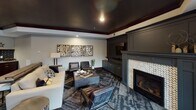 This single-floor space is thoughtfully designed as a premium entertainment and social hub, combining refined aesthetics with practical amenities across several distinct zones. The kitchenette is a standout feature, showcasing rich dark cabinetry with gold hardware, sweeping granite countertops with striking veining, and a bold deep-blue subway tile backsplash. A stainless steel sink, built-in microwave, wall oven, and refrigerator complete the setup, while industrial-style pendant lights add character overhead. Wood-look plank flooring runs throughout, adding warmth and visual continuity. The lounge area exudes an intimate, upscale ambiance with a dramatic dark ceiling framed by rich crown molding, a herringbone tile fireplace flanked by custom dark cabinetry, and a curated seating arrangement of leather chairs and a neutral sofa arranged around a coffee table. A triptych of metallic mosaic wall art adds a decorative focal point. The open game and gathering area accommodates multiple activity zones, including space for a billiards table and large communal tables, all set atop a multi-toned patterned carpet. Together, these areas create a cohesive, stylish environment well-suited for both relaxed socializing and lively entertainment.