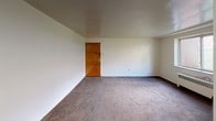This single-floor space offers a straightforward, functional layout across several connected rooms. The living area is generously sized, featuring wall-to-wall gray-toned carpeting and clean white walls that create a neutral, adaptable backdrop. A ceiling fan with an integrated light fixture provides both air circulation and overhead illumination. A solid wood interior door adds a warm contrast to the lighter surroundings. Natural light enters through a sliding window that frames a view of a grassy outdoor hillside, lending a quiet, grounded atmosphere to the space.

The 3D floor plan reveals a connected layout comprising a main living area, a secondary carpeted room, and a hallway leading to additional spaces, offering a practical flow throughout the unit.

The bathroom is compact and efficiently arranged, featuring a tub-shower combination with white surround tiling, a toilet, and a wood-cabinet vanity with an integrated sink. A small window above the tub allows natural light in while maintaining privacy. The floor is finished with vintage-style patterned tile in cream and blue tones, adding a subtle decorative element. Overall, the space presents a clean, modest, and move-in-ready environment.