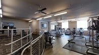 This fitness center is thoughtfully designed to accommodate a wide range of exercise needs within a single, open-plan floor. The space features durable hardwood-style flooring complemented by rubber matting in key workout zones, providing both functionality and comfort. Overhead, a ceiling fan works alongside bright fluorescent strip lighting to keep the environment well-ventilated and evenly illuminated throughout. The equipment layout is organized and spacious, with a dedicated cardio zone along the windowed wall housing treadmills and a stationary bike, allowing exercisers to enjoy natural light during their workouts. A multi-functional cable machine anchors the strength training area, while a free weight station with a dumbbell rack and adjustable bench offers versatile resistance training options. A workout bench with floor mats provides additional space for stretching and bodyweight exercises. The entrance area features a stylish curved metal railing, adding an architectural touch to the practical space. Neutral-toned walls and a framed piece of wall art lend the room a clean, welcoming ambiance. A wall-mounted television adds an entertainment element, enhancing the overall workout experience. The space is designed to be both functional and inviting for users of all fitness levels.