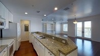 This single-floor space is defined by an open-concept layout that connects the kitchen and living areas into one cohesive, functional environment. The kitchen is a standout feature, showcasing an expansive granite countertop with speckled earth tones that extends into a generous peninsula with bar seating. White cabinetry with brushed hardware provides ample storage, while a double stainless steel sink and recessed lighting add both practicality and polish. A pendant light fixture adds a refined accent above the peninsula.

The living area flows naturally from the kitchen, offering a spacious and versatile layout anchored by rich hardwood flooring that runs throughout the space. Built-in shelving units line one wall, providing both display and storage opportunities, while a ceiling fan ensures year-round comfort. Multiple windows allow natural light to fill the room, creating a bright and welcoming ambiance. The neutral color palette of soft whites and grays throughout the space lends a clean, modern aesthetic that is both timeless and adaptable. Recessed lighting is distributed evenly across the ceiling, ensuring consistent illumination throughout. The overall design strikes a balance between practical functionality and contemporary style.