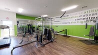 Located at 38441 Fifth St W in Palmdale, CA, this fitness center is a well-equipped, single-floor facility designed to support a wide range of workout routines. The space is defined by its bold two-tone wall design, featuring vibrant green lower panels paired with white upper sections adorned with motivational words and phrases — including "determination," "strength," "conquer," and "perseverance" — printed in varying fonts and sizes to energize members throughout their sessions.

The workout floor is finished with durable wood-style planking that extends throughout the open layout, offering both functionality and visual warmth. Full-length mirrors run along one wall, visually expanding the space and allowing users to monitor their form during exercises.

The strength training area is anchored by a multi-station cable machine with an extensive weight stack, accompanied by adjustable benches, a dumbbell rack with a full range of free weights, and a medicine ball tower. The cardio section includes treadmills, an elliptical, and an upright bike, all neatly arranged to maximize floor space. Overhead fluorescent lighting ensures the entire area is brightly and evenly lit.

An entry point connects the fitness floor to an adjacent room, providing a practical transition area. The overall ambiance is motivating and focused, making this a functional and purpose-driven fitness facility.
