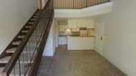 This two-story space is defined by its open, airy layout and functional design. The main level features a carpeted living area that flows openly toward the kitchen, which is positioned at the rear of the floor. The kitchen is equipped with white cabinetry — both upper and lower — offering generous storage space, along with a counter-height peninsula that provides a natural boundary between the kitchen and the living area. White appliances, including a dishwasher and refrigerator, complement the clean, neutral palette throughout. A sink is set into the countertop along the back wall, with under-cabinet lighting adding subtle illumination to the workspace. A doorway off the kitchen leads to an additional room or utility space. The staircase, positioned along the left side of the main level, features dark wood treads and a metal baluster railing, lending a contemporary industrial touch to the interior. The upper level, visible from below, overlooks the main floor through the open railing, creating a sense of vertical spaciousness and connection between the two levels. The overall ambiance is neutral and practical, with light-toned walls enhancing the brightness of the space.