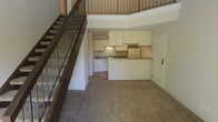 This two-story space is defined by its open, airy layout and functional design. The main level features a carpeted living area that flows openly toward the kitchen, which is positioned at the rear of the floor. The kitchen is equipped with white cabinetry — both upper and lower — offering generous storage space, along with a counter-height peninsula that provides a natural boundary between the kitchen and the living area. White appliances, including a dishwasher and refrigerator, complement the clean, neutral palette throughout. A sink is set into the countertop along the back wall, with under-cabinet lighting adding subtle illumination to the workspace. A doorway off the kitchen leads to an additional room or utility space. The staircase, positioned along the left side of the main level, features dark wood treads and a metal baluster railing, lending a contemporary industrial touch to the interior. The upper level, visible from below, overlooks the main floor through the open railing, creating a sense of vertical spaciousness and connection between the two levels. The overall ambiance is neutral and practical, with light-toned walls enhancing the brightness of the space.