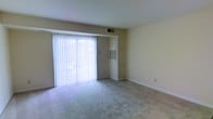 This single-floor space features a modest yet functional living area designed with simplicity and practicality in mind. The room is finished with wall-to-wall neutral carpeting that provides a warm, understated foundation throughout. A large sliding glass door dominates one wall, fitted with vertical blinds that allow for adjustable natural light control while offering access to the outdoors. The abundant natural light that filters through gives the space an open, airy feel. The walls are painted in a soft, neutral beige tone, contributing to a clean and versatile interior that can accommodate a variety of furnishing styles. An overhead HVAC vent is visible in the ceiling, and a wall-mounted utility panel is positioned near the sliding door, suggesting practical climate control options. White baseboards frame the perimeter of the room, adding a subtle contrast to the carpeted floor. The overall layout is open and unobstructed, providing flexibility in how the space can be arranged and utilized. The design is straightforward and functional, making it a practical living environment.