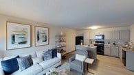 This single-floor unit offers an open-concept layout that seamlessly connects the living area and kitchen, creating a functional and inviting living environment. The living space is anchored by a comfortable neutral-toned sofa accented with navy and white throw pillows, complemented by a plush area rug and a sleek metal shelving unit used for decorative display. Framed abstract artwork adorns the walls, adding a touch of contemporary style to the warm, neutral color palette throughout. Light wood-look flooring flows consistently across the space, enhancing the sense of openness. The kitchen is equipped with black appliances, including a French door refrigerator and a range with overhead microwave, set against white cabinetry and a granite-style countertop peninsula. The peninsula doubles as a casual dining or prep space, with bar seating adding versatility. A textured ceiling adds architectural character to the space. Natural light filters in, keeping the interior bright and airy. Overall, the unit strikes a balance between practical design and modern aesthetic appeal, making it a comfortable and well-organized living space.