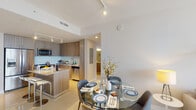 This well-appointed single-floor residence showcases a thoughtfully designed open-concept layout that seamlessly connects the kitchen, dining, and living areas. The kitchen is a standout feature, equipped with full stainless steel appliances including a French door refrigerator, built-in microwave, and range. Warm wood-toned cabinetry lines the walls and a granite-topped island with bar seating provides both additional prep space and casual dining. A glass tile backsplash adds a refined touch. The adjacent dining area accommodates a round glass-top table, while the living room benefits from an abundance of natural light streaming through floor-to-ceiling sliding glass doors that open to a private balcony overlooking the surrounding cityscape. Light-toned porcelain tile flooring runs throughout the main living areas, enhancing the bright and airy ambiance. The primary bedroom features direct balcony access, a large canvas art piece, and clean white furnishings. A second bedroom, finished in soft blue tones, includes a dedicated closet and a flexible workspace. The bathroom is outfitted with tile surround, a soaking tub and shower combination, and a granite vanity. Track lighting throughout provides versatile illumination, completing a polished, contemporary living environment.
