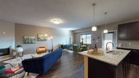 This single-floor residence is designed around an open-concept floor plan that seamlessly connects the kitchen, dining, and living areas, creating a cohesive and functional living environment. The kitchen is a standout feature, showcasing rich dark espresso cabinetry extending to the ceiling for generous storage, granite countertops, a central island with a built-in sink, stainless steel appliances, and pendant lighting overhead. Dark hardwood-style flooring runs throughout the main living areas, anchoring the space with warmth and continuity.

The living room is generously proportioned and centers around a sleek, low-profile fireplace set into the wall, providing a refined focal point. The open layout allows for distinct seating and dining zones while maintaining an airy, connected feel. Natural light filters in through windows fitted with blinds.

The bedroom features plush carpeting, a window with natural light, and comfortable proportions suitable for a full bedroom suite. The bathroom is well-appointed with dark cabinetry, a granite vanity, a large mirror, and a tub-shower combination. Throughout the space, neutral taupe wall tones and textured ceilings provide a clean, consistent backdrop.