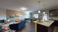 This single-floor residence is designed around an open-concept floor plan that seamlessly connects the kitchen, dining, and living areas, creating a cohesive and functional living environment. The kitchen is a standout feature, showcasing rich dark espresso cabinetry extending to the ceiling for generous storage, granite countertops, a central island with a built-in sink, stainless steel appliances, and pendant lighting overhead. Dark hardwood-style flooring runs throughout the main living areas, anchoring the space with warmth and continuity.

The living room is generously proportioned and centers around a sleek, low-profile fireplace set into the wall, providing a refined focal point. The open layout allows for distinct seating and dining zones while maintaining an airy, connected feel. Natural light filters in through windows fitted with blinds.

The bedroom features plush carpeting, a window with natural light, and comfortable proportions suitable for a full bedroom suite. The bathroom is well-appointed with dark cabinetry, a granite vanity, a large mirror, and a tub-shower combination. Throughout the space, neutral taupe wall tones and textured ceilings provide a clean, consistent backdrop.