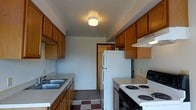 This single-floor space centers around a galley-style kitchen that maximizes functionality within a compact, efficient layout. The kitchen is lined on both sides with warm-toned wooden cabinetry, providing generous storage above and below the countertops. A double-basin sink is positioned along one wall beneath the upper cabinets, while the opposite side houses a white electric coil-top range with an overhead exhaust vent. A white refrigerator stands at the far end of the kitchen, anchoring the space and contributing to a cohesive, all-white appliance set. The countertops offer practical workspace, complemented by the abundant cabinetry surrounding them. The floor features a classic checkered vinyl tile pattern in burgundy and neutral tones, adding a touch of character to the room. A flush-mount ceiling light provides overhead illumination throughout the kitchen. The neutral gray walls keep the space feeling open and clean. A doorway at the end of the kitchen leads to additional areas of the unit, suggesting a straightforward and easy-to-navigate floor plan. Overall, the kitchen presents a no-frills, utilitarian design suited for everyday use.