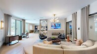 Located at 925 S Glebe Rd in Arlington, VA, this elegantly designed single-floor amenity space offers a refined and welcoming environment across three distinct areas. The open-concept lounge serves as the centerpiece, anchored by a sleek linear fireplace topped with a wall-mounted TV and flanked by a striking sculptural gold chandelier. Comfortable curved seating, leather accent chairs, and a tufted coffee table create an inviting social atmosphere, while light hardwood flooring and neutral tones lend the space a warm, contemporary feel. A built-in open shelving unit with a light wood finish acts as a thoughtful divider between the lounge and an adjacent dining or coworking area, which features dedicated seating and access to large windows that draw in natural light. The kitchenette area is outfitted with light wood cabinetry, a marble-veined waterfall island, gold pendant lighting, open shelving, and a utility sink — ideal for hosting and event use. A separate alcove features floor-to-ceiling upholstered wall panels with integrated LED cove lighting and a built-in bench, offering a cozy, intimate retreat. The cohesive design palette of navy blue accents, brass fixtures, and soft neutrals creates a polished, modern ambiance throughout.
