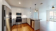 This well-designed single-floor space offers a contemporary layout with thoughtful finishes throughout. The kitchen is a standout feature, showcasing a large island topped with granite countertops and a built-in sink, complemented by a full suite of stainless steel appliances including a refrigerator, electric range, over-the-range microwave, and dishwasher. Sleek two-tone cabinetry in white and gray lines the walls, while a white subway tile backsplash and warm hardwood-style flooring add a polished, modern aesthetic. Stylish pendant lighting illuminates the kitchen and island area. The bedrooms are finished with plush wall-to-wall carpeting and feature ceiling fans, with windows that bring in natural light and outdoor views. Two bathrooms serve the unit: one features a tub-shower combination with a modern vanity and hardwood-style flooring, while the other offers a glass-enclosed shower stall, a streamlined vanity, and matching flooring. The overall ambiance is clean, bright, and contemporary, with a neutral color palette of whites and soft grays that creates a fresh and versatile living environment throughout the space.