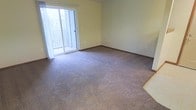 This single-floor space offers a clean, functional layout centered around a generously sized main living area. The room is finished with wall-to-wall plush carpeting in a neutral tone, providing a warm and comfortable underfoot feel throughout. Soft yellow-green walls create a calm, understated ambiance that pairs well with the abundant natural light streaming in through large sliding glass doors. These doors open directly to a private balcony, offering outdoor access and views of the surrounding greenery, further enhancing the bright and airy feel of the interior. Dark wood baseboards provide a subtle contrast to the light-colored walls, adding a touch of definition to the room's perimeter. Multiple electrical outlets are positioned along the walls, offering practical flexibility for furniture arrangement and everyday use. The open, unobstructed floor plan allows for versatile use of the space, accommodating a variety of living arrangements. The overall ambiance is quiet and neutral, making it a ready canvas for personalization. The seamless connection between the interior living area and the outdoor balcony is a standout feature, blending indoor comfort with the appeal of fresh air and natural surroundings.