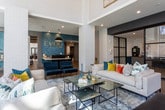 Located at 10820 Abbotts Bridge Road in Johns Creek, Georgia, this single-floor space presents a modern, design-forward interior that balances openness with intimate seating arrangements. The entrance features large black-framed glass doors and windows that flood the foyer with natural light, complemented by wide-plank hardwood flooring and white brick columns that establish a clean, contemporary aesthetic throughout.

The main lounge area is anchored by dramatic floor-to-ceiling black-framed windows offering sweeping views of the surrounding tree line, allowing abundant natural light to pour across the space. Comfortable seating arrangements—including a sofa, leather chairs, and wingback seating—are arranged around low coffee tables atop textured area rugs, creating an inviting and relaxed atmosphere. A built-in shelving unit with teal-toned accents provides display space and houses a large wall-mounted screen, adding a functional focal point to the room.

A distinct café-style nook features bold geometric black-and-white patterned tile flooring, marble-topped bistro tables, and dark bistro stools. The walls transition from white to teal wainscoting, adorned with a striking gold mosaic wood-panel artwork and a colorful canvas painting, adding an artistic, curated character to the dining area.