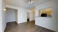 This single-floor residential space offers a clean, functional layout designed for comfortable everyday living. The main living area is open and airy, featuring dark hardwood-style flooring that extends throughout, creating a warm and cohesive aesthetic. A ceiling fan with a light fixture anchors the living space, providing both illumination and air circulation.

The kitchen is positioned along one side of the living area, separated by a partial wall with a raised counter that serves as a breakfast bar or pass-through. Light wood cabinetry and dark appliances, including a refrigerator, are visible from the living space, offering a practical and accessible cooking area.

A central hallway connects the living area to the additional rooms beyond, with a pendant light fixture illuminating the passage. The layout allows for a natural flow between spaces. The walls throughout are finished in a crisp white, contributing to a bright and neutral interior palette. The textured ceiling adds a subtle architectural detail throughout.

A doorway on the far side of the living room leads to additional spaces, suggesting a well-organized floor plan with clearly defined zones for living, dining, cooking, and private areas. Overall, the space presents a straightforward, move-in-ready environment with consistent finishes and a practical design.