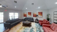 This well-appointed single-floor space offers an open and welcoming layout designed for both relaxation and social gathering. The main living area features soaring vaulted ceilings with recessed lighting and a ceiling fan, rich hardwood flooring, and a generous seating arrangement that includes leather sofas, accent chairs, and a round wooden coffee table atop a vibrant area rug. Bold abstract artwork in warm amber tones adorns the walls, adding a contemporary artistic touch throughout.

A dedicated seating nook provides a more intimate setting, furnished with charcoal and red leather chairs alongside open metal shelving displaying decorative accents. The dining area accommodates a large bar-height table and chairs, positioned adjacent to full-length French doors that flood the space with natural light and offer views of a lush outdoor greenspace.

The kitchen is thoughtfully designed with white cabinetry, dark granite countertops, a mosaic tile backsplash, and stainless steel appliances including a refrigerator, stove, microwave, and dishwasher. A teal accent wall adds a pop of color and visual interest. Tile flooring and ample counter space make this kitchen both functional and stylish. Together, these areas create a cohesive, inviting environment ideal for community use.