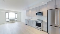 This single-floor space presents a clean, contemporary design defined by an open-concept layout that maximizes both functionality and visual flow. The kitchen is a standout feature, outfitted with floor-to-ceiling light gray flat-front cabinetry offering generous storage throughout. Stainless steel appliances — including a range with oven, over-the-range microwave, and dishwasher — complement the neutral white countertops and a simple, modern undermount sink. A white tile backsplash ties the kitchen together with a crisp, uniform aesthetic. Recessed lighting is installed throughout, providing consistent, even illumination across the space. The light-toned hardwood flooring runs seamlessly from the kitchen into the adjacent living area, reinforcing the open and airy feel of the interior. Natural light filters in through a window visible in the living area, enhancing the brightness of the space. The overall design palette — whites, grays, and natural wood tones — creates a minimalist and cohesive atmosphere that is both modern and inviting. The thoughtful layout and quality finishes make this a well-appointed, move-in ready space suited for a range of uses.
