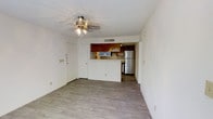 This single-floor space offers a clean, functional layout designed for comfortable everyday living. The main living area is finished with light wood-style laminate flooring that extends throughout, creating a bright and cohesive atmosphere. A ceiling fan with an integrated light fixture is centrally positioned overhead, providing both airflow and ambient lighting to the space. The living area opens toward the kitchen, which is connected via a half-wall pass-through counter — a practical design element that maintains an open feel while defining each zone. The kitchen features warm-toned wood cabinetry that adds a classic contrast to the white walls, and is equipped with a full-size refrigerator and additional appliances. A textured ceiling adds subtle visual interest throughout. The entryway, positioned near the living space, includes a solid white door with standard hardware, offering a straightforward and secure point of entry. Visible HVAC venting ensures year-round climate control. The overall palette of whites, light wood tones, and warm cabinetry gives the space a neutral, welcoming ambiance well-suited for a range of uses.