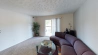 This single-floor residence offers a functional and cohesive layout across several well-defined spaces. The living room is carpeted and features a textured ceiling, neutral walls, and a sliding glass door that opens to a balcony, allowing natural light to fill the room. Adjacent to the living area, the dining room includes wood-look flooring, a ceiling fan with light fixture, wainscoting detail, and a decorative wall mirror, creating a distinct and inviting space for meals.

The galley-style kitchen is efficiently designed with rich dark wood cabinetry, granite-look countertops, a double-basin sink, and black appliances including a refrigerator, range, and microwave. Wood-look flooring continues throughout this space, complementing the cabinetry tones.

The bedroom is generously sized with plush carpet, multiple windows fitted with blinds for natural light and privacy, and a textured ceiling. A sliding closet provides ample storage, and additional dark wood furniture pieces coordinate with the room's warm tones. The bathroom features a shower with curtain, dark wood cabinetry, wood-look flooring, and hallway access connecting to the bedroom.