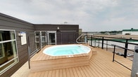 This rooftop deck offers a well-appointed outdoor retreat designed for relaxation and open-air enjoyment. The centerpiece of the space is a large octagonal hot tub, recessed into a raised platform finished in warm-toned composite decking that extends generously across the entire rooftop level. The decking material provides a clean, low-maintenance surface with a natural wood aesthetic that complements the overall design.

The deck is enclosed by sturdy black metal cable-rail fencing, ensuring safety while preserving unobstructed sightlines to the surrounding environment. From this elevated vantage point, panoramic views stretch across the surrounding landscape, including open fields and the broader urban surroundings, visible on all sides.

Architectural elements include a dark-paneled wall section featuring horizontal louvered panels, adding a modern, finished look to the rooftop structure. Exterior lighting fixtures are mounted on the building facade, supporting use of the space during evening hours. Access to the hot tub is facilitated by built-in steps with handrails on either side.

Overall, this rooftop deck creates an inviting, resort-like atmosphere with a focus on open sky exposure, far-reaching views, and premium amenities — making it a distinctive and functional outdoor amenity space.