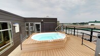 This rooftop deck offers a well-appointed outdoor retreat designed for relaxation and open-air enjoyment. The centerpiece of the space is a large octagonal hot tub, recessed into a raised platform finished in warm-toned composite decking that extends generously across the entire rooftop level. The decking material provides a clean, low-maintenance surface with a natural wood aesthetic that complements the overall design.

The deck is enclosed by sturdy black metal cable-rail fencing, ensuring safety while preserving unobstructed sightlines to the surrounding environment. From this elevated vantage point, panoramic views stretch across the surrounding landscape, including open fields and the broader urban surroundings, visible on all sides.

Architectural elements include a dark-paneled wall section featuring horizontal louvered panels, adding a modern, finished look to the rooftop structure. Exterior lighting fixtures are mounted on the building facade, supporting use of the space during evening hours. Access to the hot tub is facilitated by built-in steps with handrails on either side.

Overall, this rooftop deck creates an inviting, resort-like atmosphere with a focus on open sky exposure, far-reaching views, and premium amenities — making it a distinctive and functional outdoor amenity space.