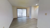 This two-story space presents a clean and functional layout with a neutral, modern aesthetic throughout. The main living area is finished with light wood-style luxury vinyl plank flooring that runs seamlessly across the floor, creating a cohesive and easy-to-maintain surface. Crisp white walls and white baseboards contribute to a bright, airy atmosphere that feels open and welcoming.

A large sliding glass door, dressed with vertical blinds, serves as a prominent feature of the living area, allowing natural light to filter into the room while providing direct access to an outdoor space. Recessed lighting in the ceiling complements the natural light, ensuring the interior remains well-lit at any time of day. The textured ceiling adds a subtle architectural detail to the otherwise minimalist space.

A structural column and partial wall define the transition between the living area and the staircase, which leads to the second floor. This layout creates a natural sense of separation and flow within the open plan. Overall, the space offers a tidy, move-in-ready environment with a simple, contemporary feel suited to a variety of lifestyles.