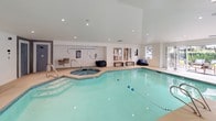 Located at 12224 Daphne Ln NW in Silverdale, Washington, this single-level indoor pool facility offers a well-appointed aquatic and relaxation space designed for year-round use. The centerpiece is a generously sized indoor swimming pool with lane markers, complemented by an adjoining octagonal spa/hot tub, both finished with light blue tile work that enhances the bright, clean aesthetic of the space.

The interior is characterized by crisp white walls, recessed lighting, and smooth concrete-style flooring that extends throughout. Large windows and multiple glass-panel doors flood the space with natural light while framing views of the surrounding mature landscaping and outdoor patio areas. Several door access points connect the interior seamlessly to exterior lounge spaces, creating a fluid indoor-outdoor experience.

A dedicated lounge area features a circular upholstered daybed with an array of decorative pillows, accent chairs, and casual seating, providing a comfortable retreat alongside the pool. Colorful artwork adorns the walls in a secondary seating nook near what appears to be a sauna or changing room access. Safety equipment, including a life ring and fire extinguisher, is visibly and conveniently placed throughout the space. The overall ambiance is modern, airy, and resort-inspired.