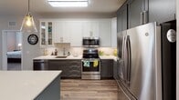 This thoughtfully designed single-floor residence in Dresher, PA offers a cohesive blend of modern finishes and functional living spaces. The kitchen is a standout feature, showcasing two-tone cabinetry with charcoal lower cabinets and crisp white uppers, a herringbone tile backsplash, light quartz countertops, a large center island, and a full suite of stainless steel appliances, including a French door refrigerator, electric range, and built-in microwave. A skylight overhead fills the kitchen with natural light. Wood-look flooring extends through the main living areas, connecting the kitchen seamlessly to the living room, which features large windows and a glass door that invites natural light and outdoor views. The primary bedroom is generously sized with plush carpeting, crown molding, a ceiling fan, and direct access to a spa-inspired en-suite bathroom complete with a floating double-sink vanity, framed mirrors, modern sconce lighting, and a glass-enclosed marble-tiled shower. A second bedroom offers comfortable proportions, its own ceiling fan, crown molding, and a bright window with exterior views. The secondary bathroom features marble-tiled walls, a soaking tub with shower, and clean white fixtures, maintaining the home's refined, contemporary aesthetic throughout.