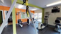 This fitness center is designed to support a well-rounded workout routine in a bright and energizing environment. The space is finished with durable rubber flooring suitable for both cardio and strength training activities. Bold accent walls in vibrant orange and lime green lend the room an energetic, motivating atmosphere, complemented by crisp white trim and ample overhead lighting.

The facility is equipped with a multi-station cable weight machine, TRX suspension training straps mounted to an overhead anchor, and a selection of cardio equipment including an elliptical trainer and a recumbent-style cycle. A large mirror is mounted along one wall, enhancing the sense of space and allowing users to monitor their form during exercise.

Natural light fills the room through multiple large windows, which also provide pleasant views of the surrounding outdoor landscaping, including colorful plantings and greenery beyond a perimeter fence. A wall-mounted HVAC unit ensures year-round climate comfort during workouts.

The overall layout is thoughtfully organized to allow users to move freely between equipment, making the space functional for both individual training sessions and varied fitness routines. The single-floor layout ensures easy accessibility throughout the facility.