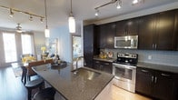 This contemporary single-floor residence offers a thoughtfully designed open-concept layout that seamlessly connects the kitchen, dining, and living areas. The kitchen serves as the centerpiece of the space, showcasing rich dark espresso cabinetry that extends to the ceiling for ample storage, complemented by granite countertops with a deep undermount stainless steel sink. A large kitchen island anchors the space, doubling as a breakfast bar and entertaining hub. Stainless steel appliances, including an electric range with oven and an over-the-range microwave, add a sleek, modern touch, while a glass tile backsplash provides a refined finishing detail. Track lighting and elegant pendant lights illuminate the kitchen and dining area, creating a warm and inviting ambiance. The dining area comfortably accommodates a full table setting, ideal for entertaining. Light hardwood flooring flows throughout, brightening the interior. A glimpse into the adjacent bedroom reveals a bright, well-appointed sleeping space, highlighting the cohesive and polished design aesthetic that runs throughout this well-appointed residence.
