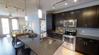 This contemporary single-floor residence offers a thoughtfully designed open-concept layout that seamlessly connects the kitchen, dining, and living areas. The kitchen serves as the centerpiece of the space, showcasing rich dark espresso cabinetry that extends to the ceiling for ample storage, complemented by granite countertops with a deep undermount stainless steel sink. A large kitchen island anchors the space, doubling as a breakfast bar and entertaining hub. Stainless steel appliances, including an electric range with oven and an over-the-range microwave, add a sleek, modern touch, while a glass tile backsplash provides a refined finishing detail. Track lighting and elegant pendant lights illuminate the kitchen and dining area, creating a warm and inviting ambiance. The dining area comfortably accommodates a full table setting, ideal for entertaining. Light hardwood flooring flows throughout, brightening the interior. A glimpse into the adjacent bedroom reveals a bright, well-appointed sleeping space, highlighting the cohesive and polished design aesthetic that runs throughout this well-appointed residence.