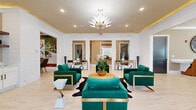 Located at 9311 Pine Forest Road in Pensacola, Florida, this single-floor space delivers a striking blend of contemporary elegance and functional design across a well-appointed layout. The living room serves as the centerpiece, featuring light marble-look tile flooring, a tray ceiling with warm gold accent lighting, and a dramatic botanical chandelier. Emerald green velvet seating with gold-trimmed frames creates a bold, sophisticated focal point, complemented by a cowhide area rug and abundant natural light streaming through large windows flanking the entrance doors. A built-in beverage station with white cabinetry and open shelving adds a convenient, polished touch to the living area. The kitchen is thoughtfully equipped with white shaker-style cabinetry, a decorative geometric tile backsplash, stainless steel appliances, and generous counter space, all illuminated by a window that brings in natural light. Crown molding throughout enhances the refined character of the space. The hallway connects the rooms seamlessly, featuring neutral warm-toned walls, clean tile flooring, and a large-scale abstract artwork in a gold frame that adds an artistic statement. The dining area rounds out the layout, offering an intimate setting ideal for gatherings. Overall, the space exudes a polished, upscale ambiance with cohesive design details from room to room.