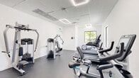 Located at 25850 SW 140th Ct in Homestead, FL, this fitness center offers a clean, modern exercise environment within a single, well-organized room. The space features crisp white walls, a drop-panel ceiling with recessed linear LED lighting, and durable dark rubber flooring designed to withstand heavy equipment use.

The cardio section is equipped with multiple treadmills and stationary bikes, including recumbent and upright models, several of which feature integrated digital displays. Strength training needs are addressed by a multi-station cable machine, a chest press machine, and an adjustable workout bench. A dedicated free weights area includes a rack of colorful medicine balls and a tiered dumbbell rack, providing options for a full range of resistance training. A wall-mounted mirror near the strength zone enhances the sense of space and supports proper form.

Natural light filters in through large windows on one wall, offering views of palm trees and the surrounding community — a bright, Florida-inspired atmosphere that keeps the space feeling open and inviting. Ventilation is supported by ceiling-mounted vents and a wall unit, ensuring comfort during workouts. The layout is efficient and thoughtfully arranged, making the most of the available floor area while maintaining clear pathways between equipment stations. Signage and safety notices are posted throughout, reflecting an attentive, well-maintained facility.