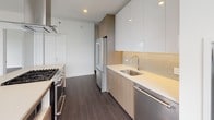 This thoughtfully designed single-floor residence showcases a contemporary aesthetic throughout, combining clean lines, natural wood tones, and a crisp white palette. The open-concept kitchen serves as the centerpiece, anchored by a substantial island with an integrated gas cooktop and a stainless steel range hood above. Cabinetry blends high-gloss white uppers with warm wood-grain lowers, complemented by light subway tile backsplash and sleek quartz countertops. Stainless steel appliances, including a French door refrigerator and dishwasher, complete the well-appointed space. Dark hardwood flooring flows consistently throughout the living areas and bedrooms, providing a grounding contrast to the bright white walls and ceilings. Two bedrooms each feature built-in closet systems with shelving and hanging rods, offering generous storage. The primary bathroom includes a frameless glass walk-in shower clad in large-format neutral tiles, a wood-paneled vanity with an undermount sink, and a large mirror. A secondary bathroom features similar wood-toned cabinetry and light tile flooring. Floor-to-ceiling windows in the living area and bedroom flood the space with natural light while framing expansive views of the surrounding cityscape and treetops.