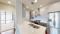 This thoughtfully designed 2-bedroom, 2-bathroom residence offers a cohesive modern aesthetic throughout its single-floor layout. The kitchen serves as a standout centerpiece, featuring quartz countertops with a double stainless steel sink, gray shaker-style cabinetry, a subway tile backsplash, stainless steel appliances including a gas range and French door refrigerator, a professional-grade range hood, and a pendant light accent — all combining for a sleek, functional workspace. The living room is bathed in natural light through large windows framing urban cityscape views, and features warm hardwood-style flooring and a ceiling fan overhead. Both bedrooms are carpeted for comfort, each offering generous windows with city views and ceiling fans. The primary bedroom includes a dedicated closet with built-in shelving for organized storage. The two bathrooms are finished consistently with gray cabinetry, quartz countertops, and light tile flooring. One bathroom features a tub-shower combination with classic subway tile surround, while the other offers a single vanity with a large mirror. The space also includes two closets, providing practical storage solutions throughout. Clean neutral walls and recessed lighting tie the entire residence together with a polished, contemporary feel.