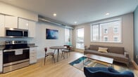 This thoughtfully designed single-floor residence showcases a contemporary industrial aesthetic, highlighted by exposed cylindrical ductwork that runs through the main living areas and bedroom, adding a distinctive urban character to the space. Light hardwood flooring flows seamlessly throughout, unifying each area with warmth and continuity.

The open-concept main living area combines the living room, dining space, and kitchen into one cohesive zone. The living room is anchored by large windows fitted with horizontal blinds that invite generous natural light and offer views of the surrounding neighborhood. The kitchen features sleek white cabinetry, granite countertops, and a full suite of stainless steel appliances, including a refrigerator, dishwasher, range, and microwave. An accent wall in deep teal adds a bold contrast to the otherwise neutral palette.

The bedroom continues the industrial-modern aesthetic with exposed ductwork overhead, recessed lighting, and a serene, neutral color scheme that creates a restful atmosphere. The bathroom is clean and well-appointed, featuring granite countertops on the vanity, large-format tile flooring, and a full tub-shower combination with white subway tile surround. Together, these spaces form a cohesive, stylish residence with a practical and modern sensibility.