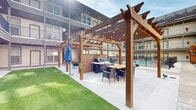 Located at 203 S Riverfront Dr in Jenks, Oklahoma, this outdoor courtyard amenity space is designed to foster community and relaxation in a resort-inspired setting. The centerpiece of the space is a sparkling swimming pool, enclosed by wrought iron fencing and surrounded by a generous pool deck outfitted with lounge chairs and seating — ideal for unwinding in the Oklahoma sunshine.

Flanking the pool area are two striking wood pergola structures, each accented with warm string lights that lend an inviting ambiance during evening hours. One pergola shelters a dedicated grilling station built into a brick enclosure, paired with outdoor dining furniture, making it well-suited for gatherings and cookouts. A well-maintained stretch of turf lawn provides additional open recreational space adjacent to the pergola area.

The courtyard is framed on multiple sides by a multi-story building featuring horizontal lap siding in a neutral palette, individual unit balconies, and wide windows that overlook the shared amenity space below. Covered walkways with recessed lighting line the ground-level perimeter, providing sheltered access to individual unit entries. Decorative wooden privacy screens and gravel landscaping add architectural texture along the building's exterior, completing a thoughtfully designed outdoor environment.