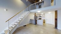 This two-story loft-style space blends industrial character with modern finishes to create a striking and functional interior. The main level features polished concrete floors that extend throughout the open-plan living area, anchored by exposed rustic wood columns that add warmth and architectural interest. Soaring ceilings amplify the sense of volume, while a white metal staircase with a wood handrail connects the two levels, serving as both a functional and visual centerpiece.

The kitchen is thoughtfully designed and positioned at the heart of the main floor. It features a large island with a granite countertop and an integrated sink, complemented by light wood cabinetry and a classic white subway tile backsplash. Stainless steel appliances, including a refrigerator, gas range, and microwave, outfit the space for full functionality. Pendant lighting above the island and flush-mount ceiling fixtures provide layered illumination throughout. A stacked washer and dryer unit is conveniently tucked into a dedicated alcove nearby.

The upper level, accessible via the staircase, overlooks the main living area through an open railing, reinforcing the loft's open and airy character. The combination of raw industrial elements and clean contemporary finishes gives this space a distinctive, versatile aesthetic.