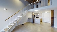 This two-story loft-style space blends industrial character with modern finishes to create a striking and functional interior. The main level features polished concrete floors that extend throughout the open-plan living area, anchored by exposed rustic wood columns that add warmth and architectural interest. Soaring ceilings amplify the sense of volume, while a white metal staircase with a wood handrail connects the two levels, serving as both a functional and visual centerpiece.

The kitchen is thoughtfully designed and positioned at the heart of the main floor. It features a large island with a granite countertop and an integrated sink, complemented by light wood cabinetry and a classic white subway tile backsplash. Stainless steel appliances, including a refrigerator, gas range, and microwave, outfit the space for full functionality. Pendant lighting above the island and flush-mount ceiling fixtures provide layered illumination throughout. A stacked washer and dryer unit is conveniently tucked into a dedicated alcove nearby.

The upper level, accessible via the staircase, overlooks the main living area through an open railing, reinforcing the loft's open and airy character. The combination of raw industrial elements and clean contemporary finishes gives this space a distinctive, versatile aesthetic.