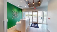 The lobby at 2424 Tulane offers a clean, contemporary first impression defined by a well-considered blend of modern design elements. At the heart of the entry area sits a crisp white reception desk, positioned in front of a vibrant green accent wall adorned with a striking abstract sculptural installation composed of intersecting wooden elements. Large floor-to-ceiling windows and glass entry doors flood the reception zone with natural light, providing an open, welcoming atmosphere and clear views to the exterior. A branded entryway mat reinforces the building's identity at the threshold.

The polished concrete floors extend throughout, lending an industrial-chic quality that ties the entire space together. Overhead, a recessed ceiling tray features a cluster of pendant lights with cylindrical fixtures, adding warmth and visual interest. Beyond the reception area, a wide corridor leads deeper into the building, lined with smooth gray doors providing access to additional rooms and facilities, including ADA-accessible restrooms. The hallway is accented with petite round pedestal tables and a bench seating arrangement, alongside curated wall art that contributes to the gallery-like ambiance. The overall design is bright, minimalist, and professional, creating an efficient and polished environment suited for a modern commercial or medical office setting.