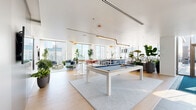 Located in Chicago's vibrant West Loop neighborhood at 123 N Desplaines St, this stylish amenity lounge offers a generously proportioned, open-plan layout flooded with natural light from expansive floor-to-ceiling windows that frame sweeping views of the city skyline. The light oak hardwood flooring and crisp white ceilings create a bright, airy atmosphere throughout the space.

The main area is anchored by a sleek pool table atop a soft area rug, surrounded by high-top bar tables and modern pedestal stools — ideal for social gatherings. Adjacent to this is a well-appointed bar counter with a bold navy blue island and white bar stools, positioned alongside full-height glass walls that provide direct access to a furnished outdoor terrace.

Multiple distinct lounge zones feature contemporary, modular seating in soft blue, green, and neutral tones, accented with colorful throw pillows and lush indoor greenery. A more intimate seating area with a round wood dining table and mint green chairs offers a relaxed setting for smaller groups.

Floor-to-ceiling glass partitions divide portions of the space while maintaining visual openness. Wall-mounted flat-screen televisions, pendant lighting with warm gold accents, and abundant tropical plants contribute to a polished yet welcoming ambiance throughout this exceptional urban retreat.